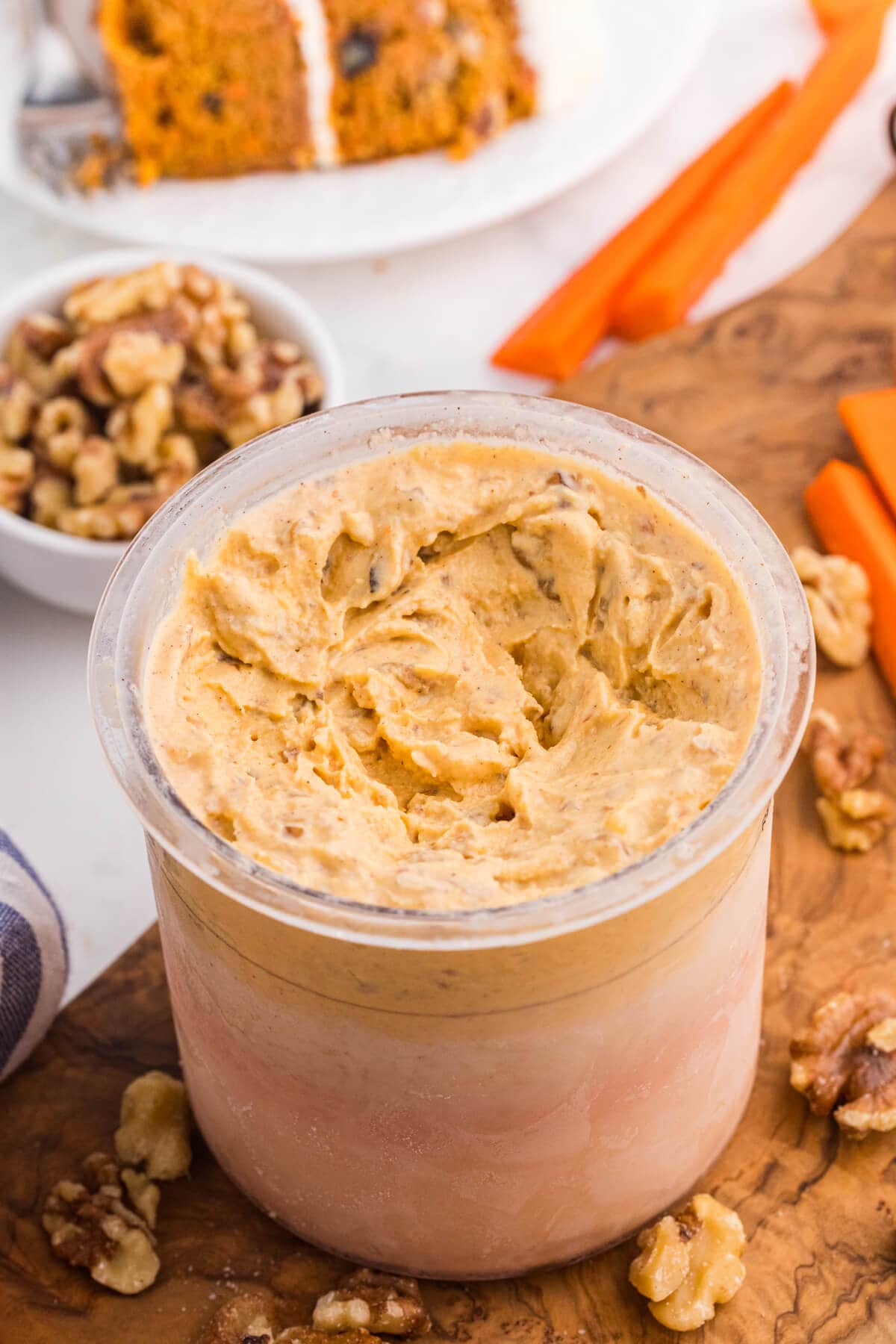 Carrot cake ice cream in a Ninja Creami pint with chopped walnuts, carrot sticks, and carrot cake in the background.