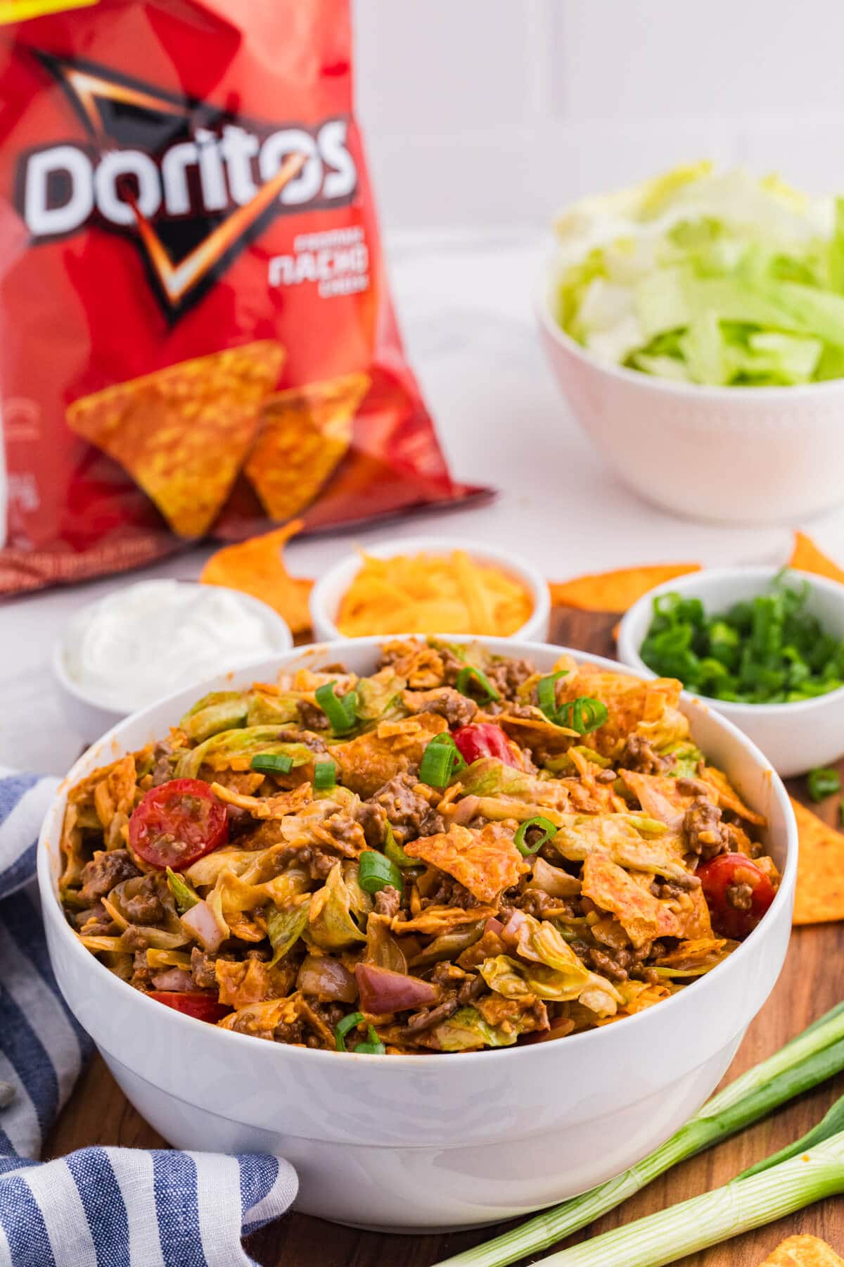 Dorito Taco Salad in a white bowl with seasoned beef, lettuce, tomatoes, red onion, cheddar cheese, green onions, and crushed Doritos, with a bag of Doritos in the background.