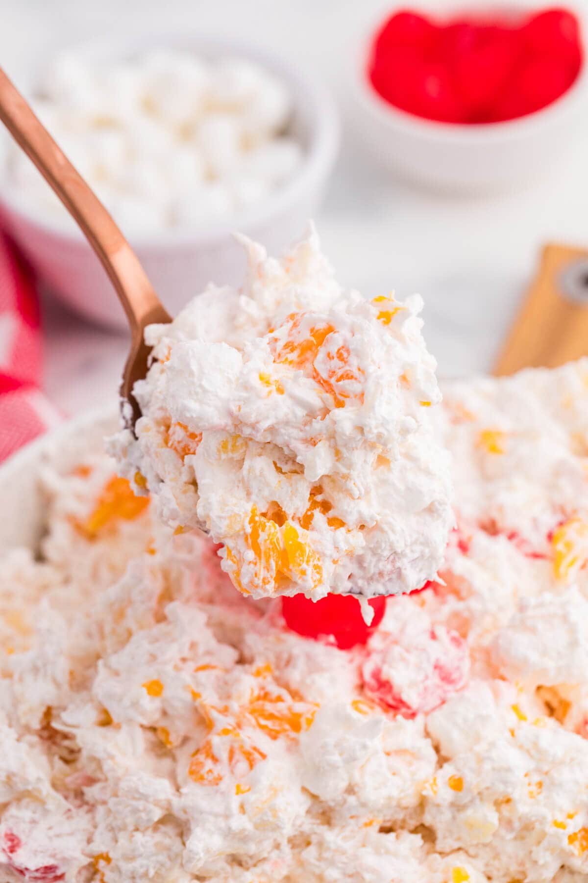A copper spoon lifts a creamy scoop of glorified rice filled with mandarin orange pieces and mini marshmallows. The remaining fruit salad with bright red maraschino cherries fills the background.