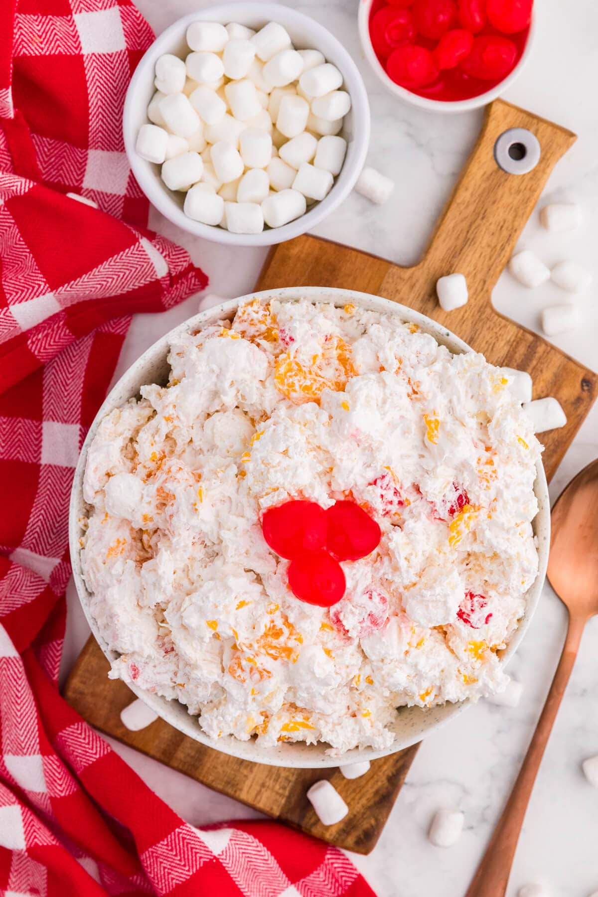 A bowl of creamy glorified rice mixed with mini marshmallows and orange and red fruit pieces. Three bright red cherries top the dessert beside bowls of extra marshmallows and cherries on a wooden board with a red plaid cloth.