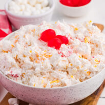 A speckled bowl of creamy glorified rice filled with mandarin oranges crushed pineapple mini marshmallows and maraschino cherries sits on a wooden board. Three bright red cherries top the dessert beside a red plaid cloth and copper spoon.