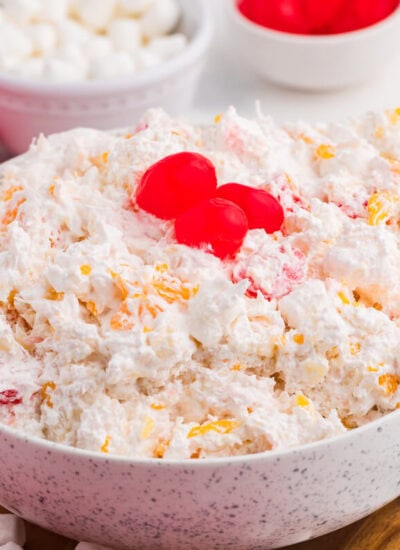 A speckled bowl of creamy glorified rice filled with mandarin oranges crushed pineapple mini marshmallows and maraschino cherries sits on a wooden board. Three bright red cherries top the dessert beside a red plaid cloth and copper spoon.