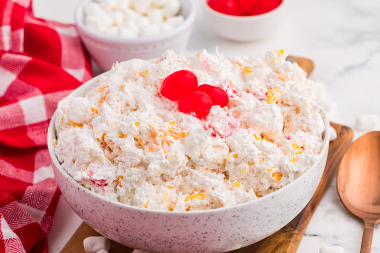 A speckled bowl of creamy glorified rice filled with mandarin oranges crushed pineapple mini marshmallows and maraschino cherries sits on a wooden board. Three bright red cherries top the dessert beside a red plaid cloth and copper spoon.