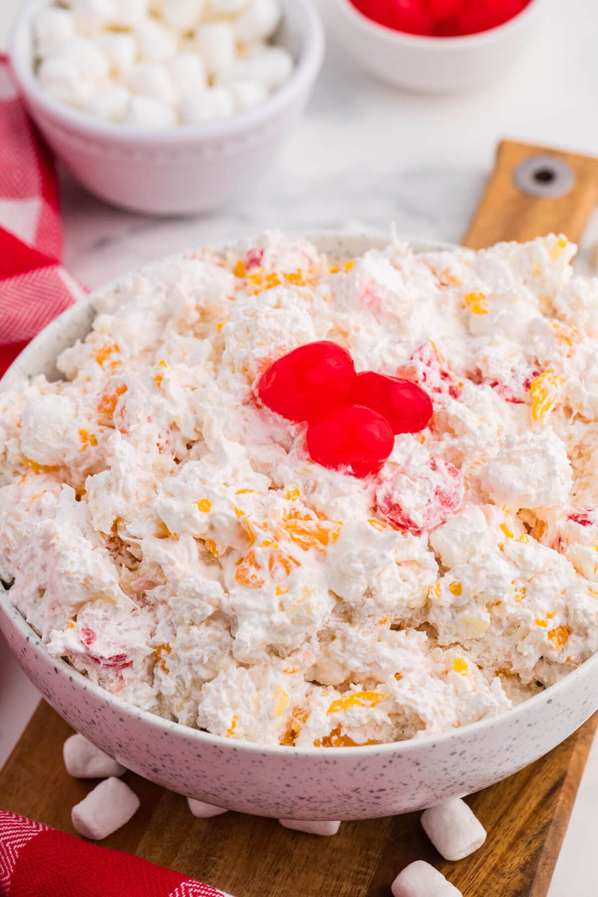 A close up of creamy glorified rice with mandarin oranges and maraschino cherries mixed throughout. Three bright red cherries top the finished dessert with mini marshmallows scattered around the bowl.