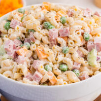 A white bowl is filled with creamy ham and pea pasta salad made with elbow macaroni diced ham green peas cheddar cubes celery and black pepper. A blue gingham cloth and blurred bowl of cheddar sit nearby for a casual serving scene.