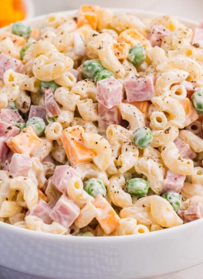 A white bowl is filled with creamy ham and pea pasta salad made with elbow macaroni diced ham green peas cheddar cubes celery and black pepper. A blue gingham cloth and blurred bowl of cheddar sit nearby for a casual serving scene.