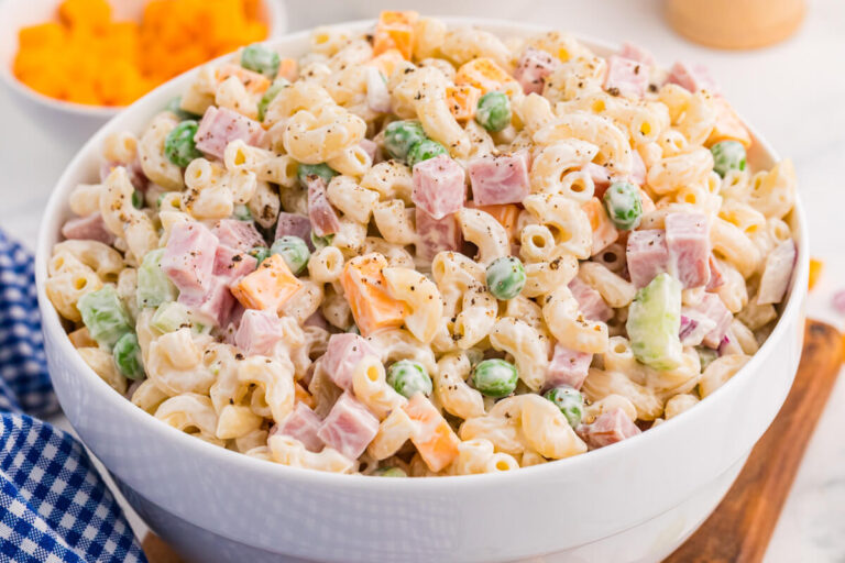 A white bowl is filled with creamy ham and pea pasta salad made with elbow macaroni diced ham green peas cheddar cubes celery and black pepper. A blue gingham cloth and blurred bowl of cheddar sit nearby for a casual serving scene.