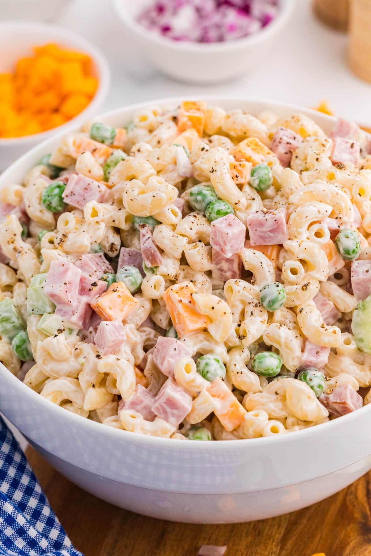 Close up bowl of creamy ham and pea pasta salad with elbow macaroni diced ham green peas cheddar cubes celery and black pepper. A blue gingham cloth and blurred bowls of cheddar and red onion frame the finished pasta salad for serving.