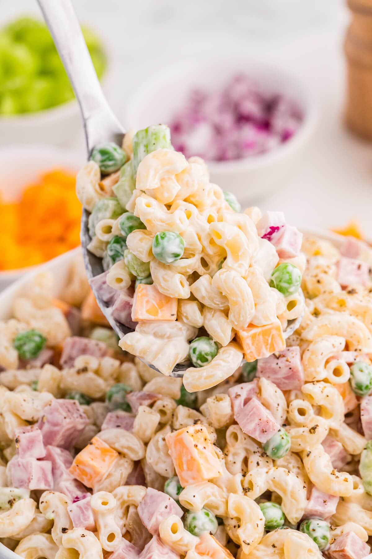 A spoon lifts creamy ham and pea pasta salad from a bowl with elbow macaroni diced ham green peas cheddar cubes celery and black pepper. Blurred bowls of cheddar celery and red onion sit in the background to show the finished pasta salad texture.