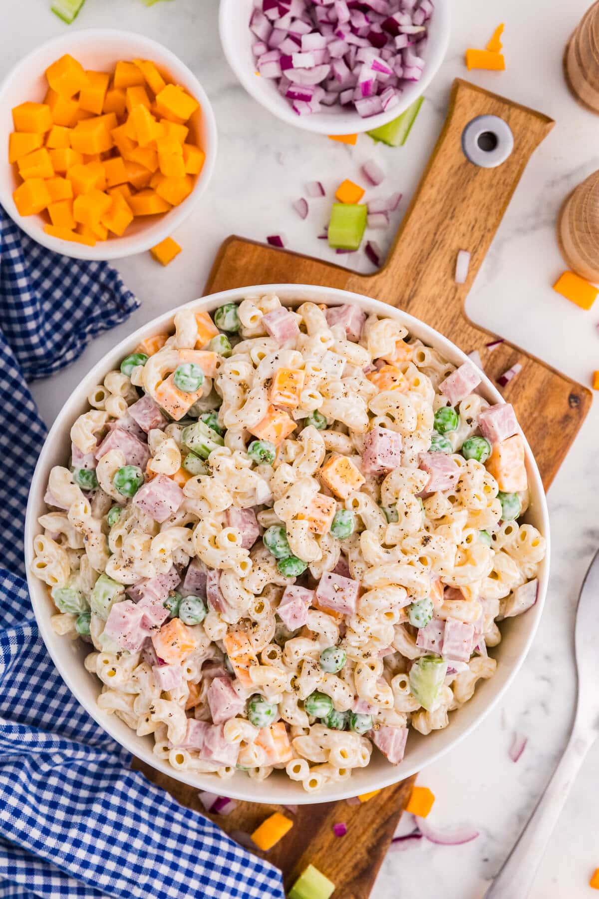 Creamy ham and pea pasta salad fills a white bowl with elbow macaroni diced ham green peas cheddar cubes celery and black pepper. Bowls of chopped cheddar and red onion sit nearby on a marble countertop with a blue gingham cloth.
