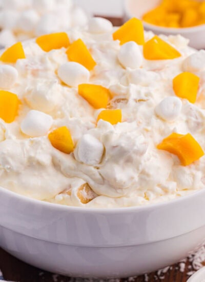 Peach fluff salad in a white bowl topped with chopped peaches and mini marshmallows, served with coconut and extra marshmallows on a wooden board.