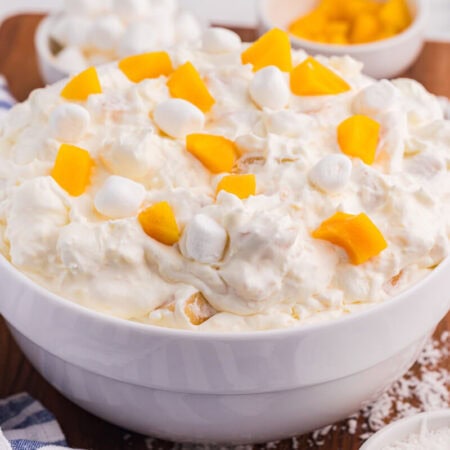 Peach fluff salad in a white bowl topped with chopped peaches and mini marshmallows, served with coconut and extra marshmallows on a wooden board.