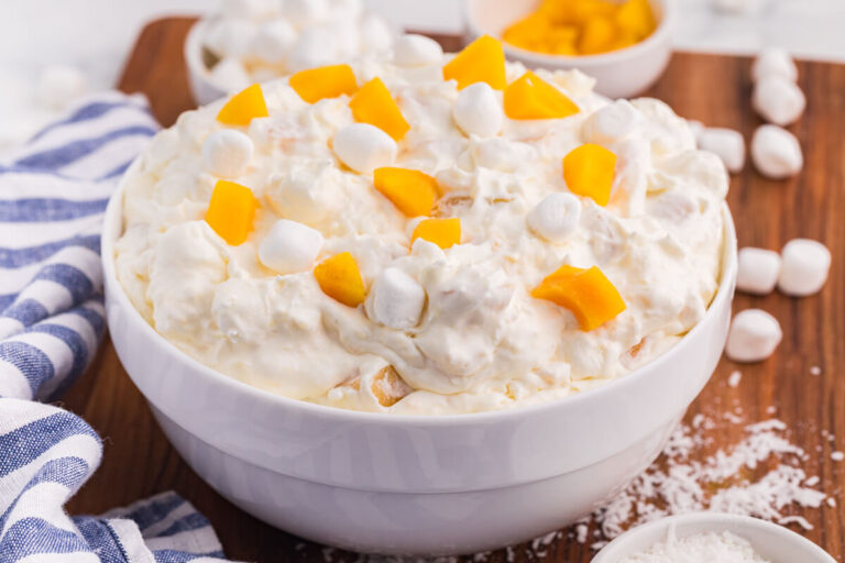 Peach fluff salad in a white bowl topped with chopped peaches and mini marshmallows, served with coconut and extra marshmallows on a wooden board.