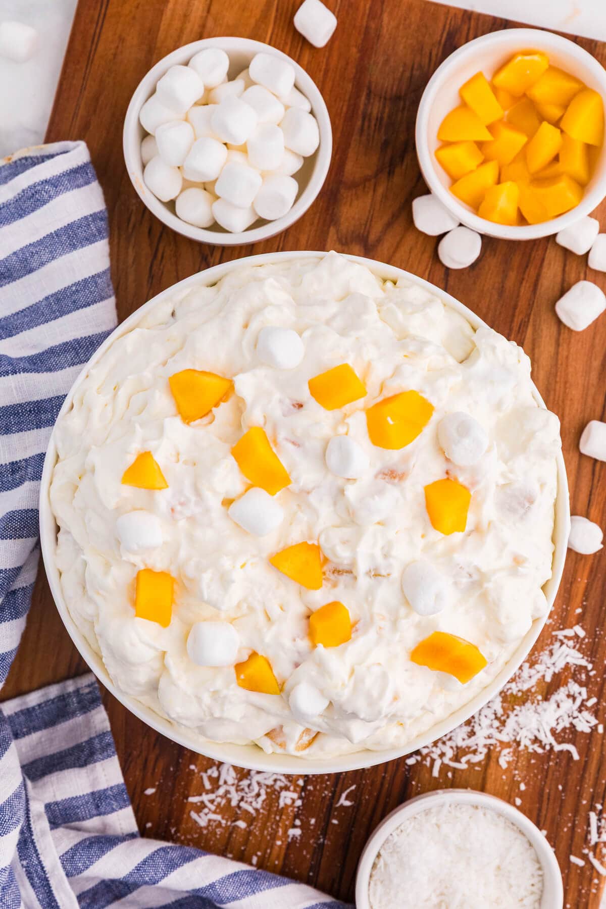 Peach fluff salad in a white bowl topped with chopped peaches and mini marshmallows, with bowls of marshmallows, peaches, and coconut on a wooden board.
