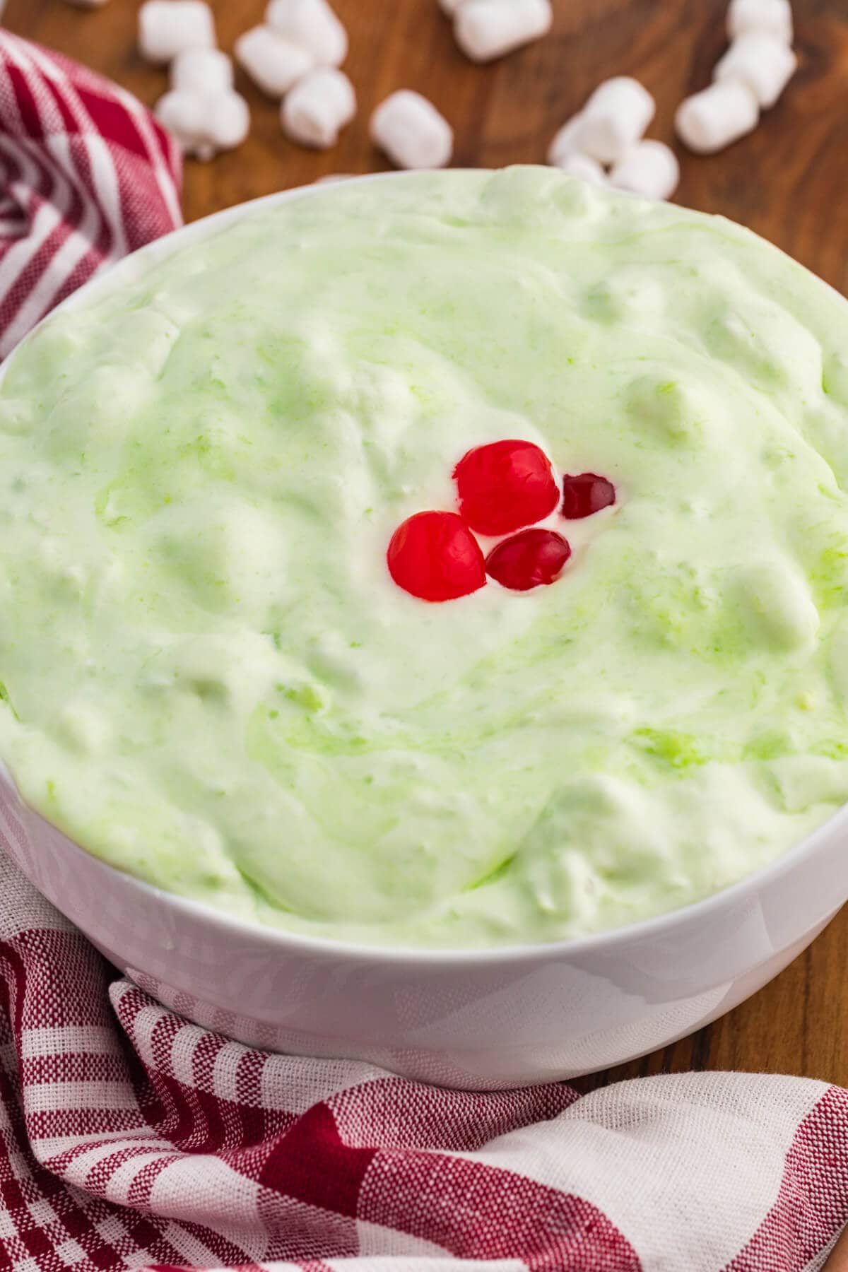Close up bowl of pale green sea foam salad topped with bright red cherries. Mini marshmallows sit in the background with a red and white checkered towel around the bowl.