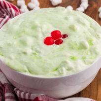 White bowl of pale green sea foam salad topped with red cherries on a wooden table. Mini marshmallows. A red and white checkered towel. And a copper spoon sit around the bowl.