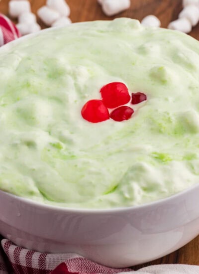 White bowl of pale green sea foam salad topped with red cherries on a wooden table. Mini marshmallows. A red and white checkered towel. And a copper spoon sit around the bowl.