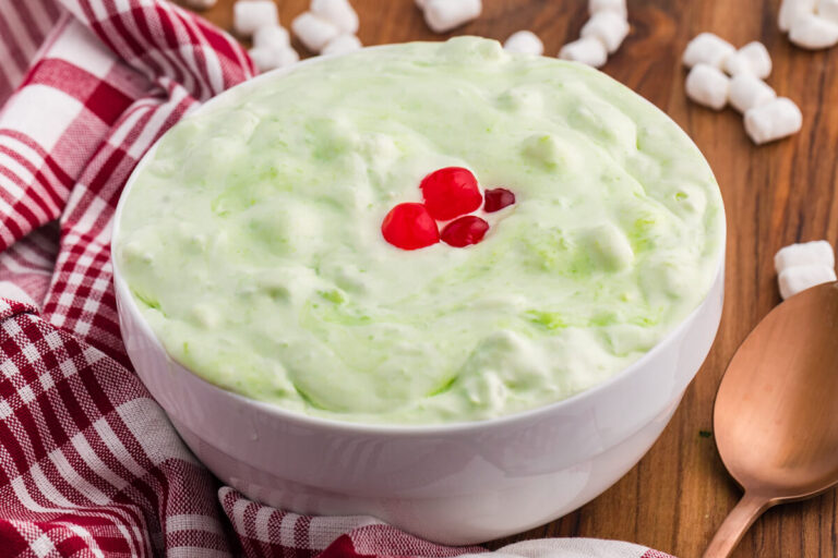 White bowl of pale green sea foam salad topped with red cherries on a wooden table. Mini marshmallows. A red and white checkered towel. And a copper spoon sit around the bowl.