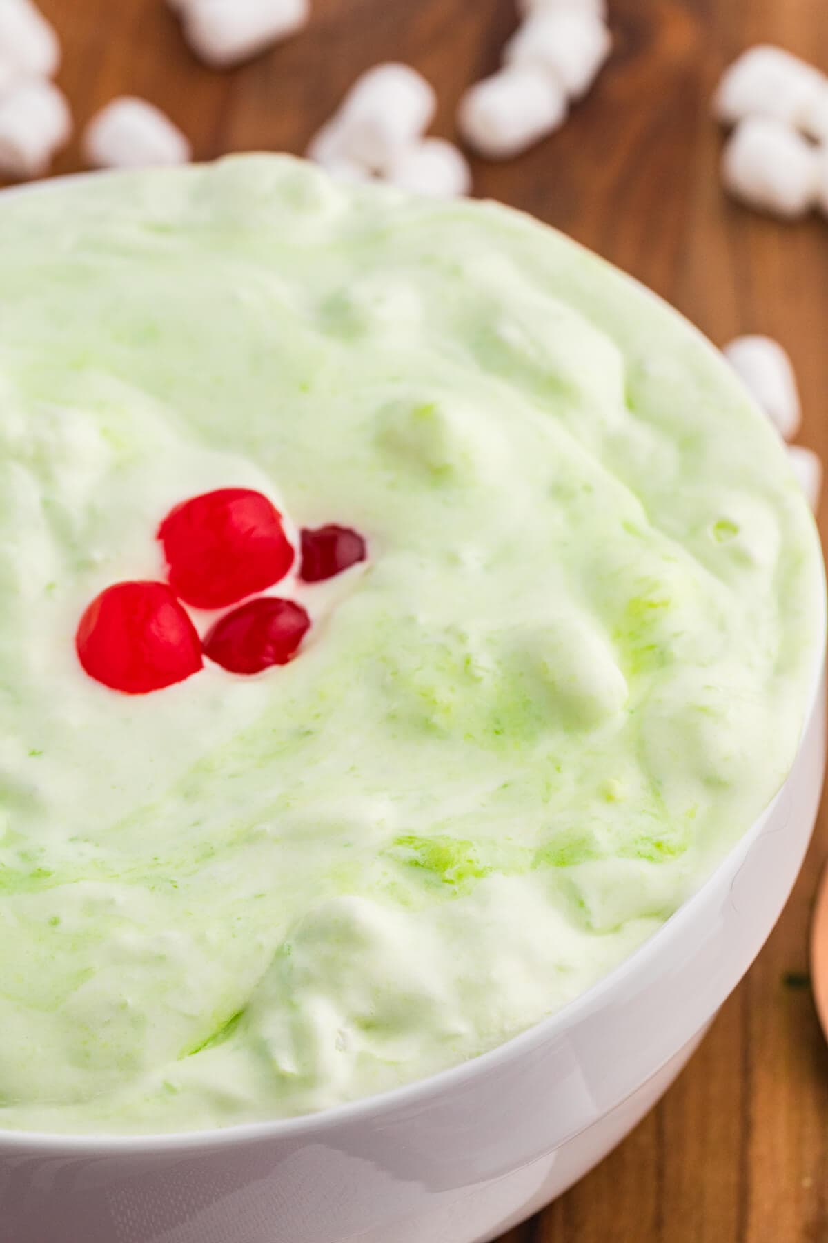 Close up of pale green sea foam salad in a white bowl topped with red cherries. Mini marshmallows and a copper spoon are blurred on the wooden table in the background.