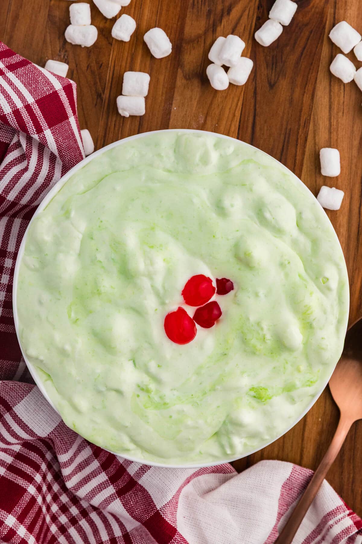 Bowl of pale green sea foam salad topped with red cherries on a wooden table. Mini marshmallows are scattered around the bowl with a red and white checkered towel and copper spoon nearby.