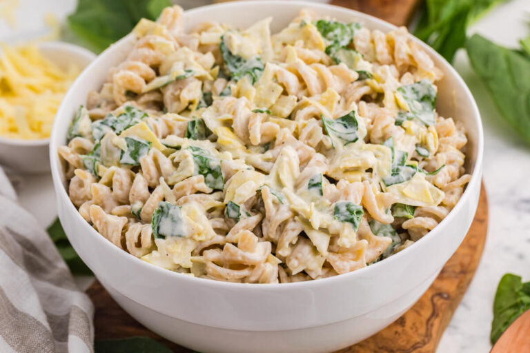 Creamy spinach artichoke pasta salad with rotini, spinach, artichokes, and cheese in a white serving bowl.