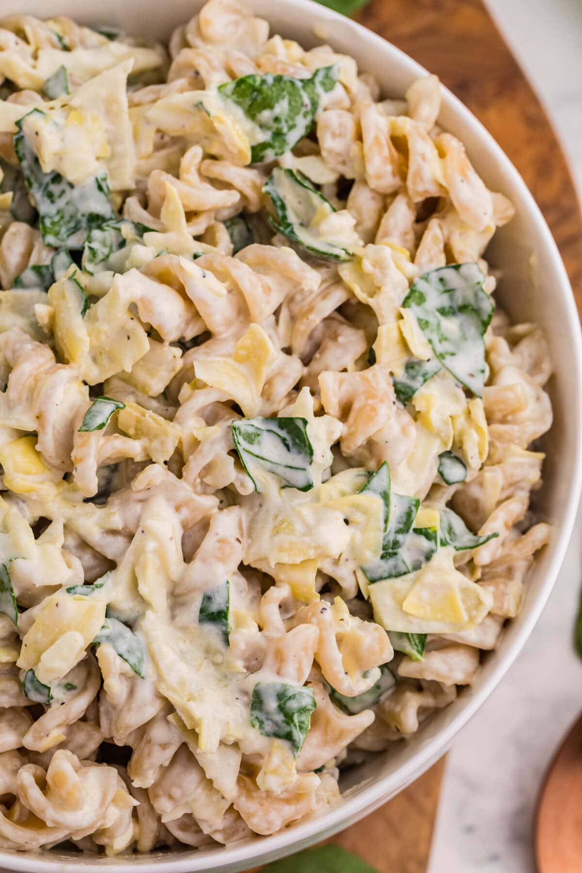 Close-up of creamy spinach artichoke pasta salad with rotini, chopped spinach, artichokes, and cheese in a white bowl.