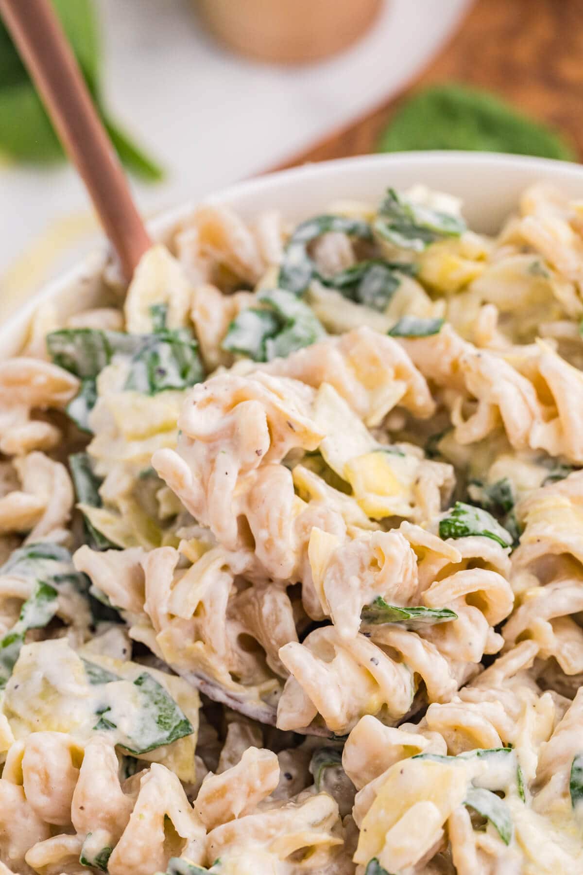 Creamy spinach artichoke pasta salad with rotini, chopped spinach, artichokes, and cheese being scooped from a bowl.