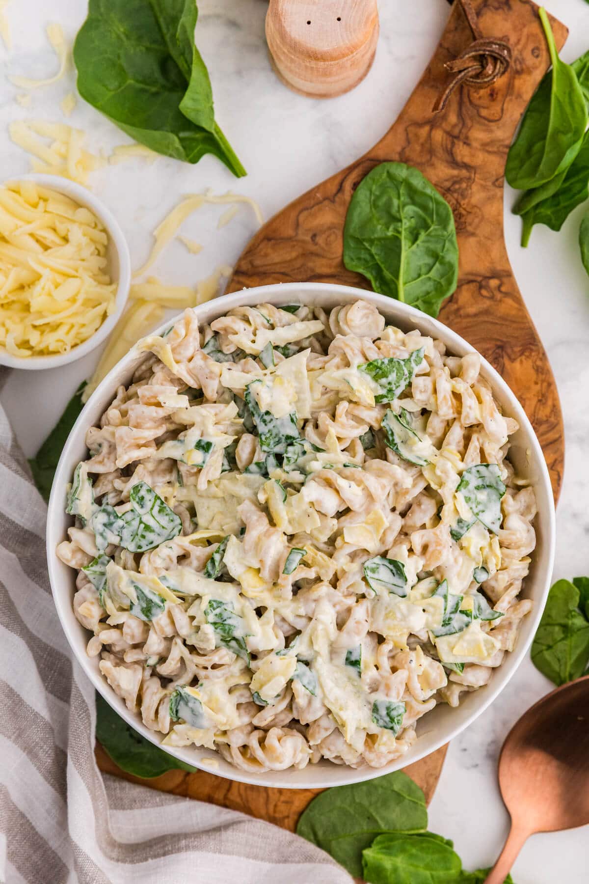Creamy spinach artichoke pasta salad in a white bowl with fresh spinach, shredded cheese, and a wooden serving spoon around it.