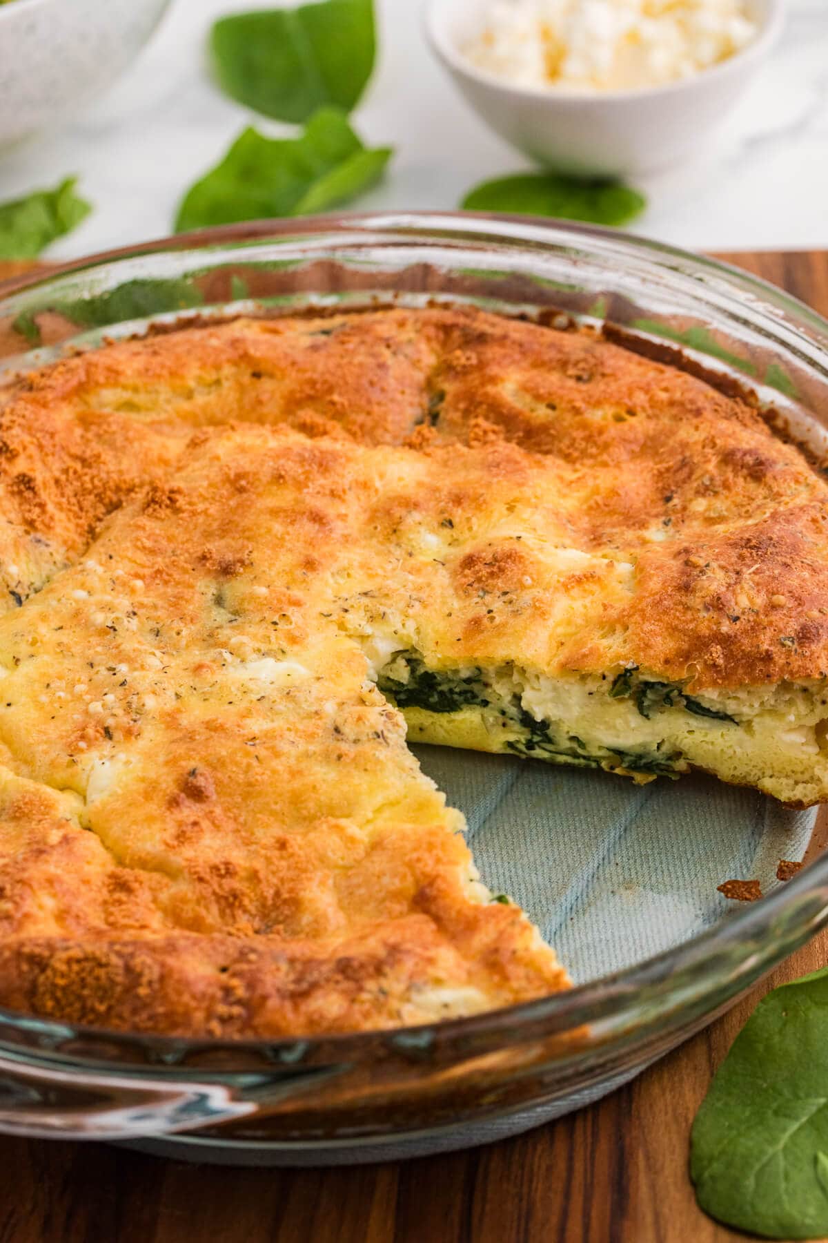 Spinach and feta impossible pie baked in a glass pie dish with a slice removed to show the creamy filling with spinach and cheese under a golden browned top.