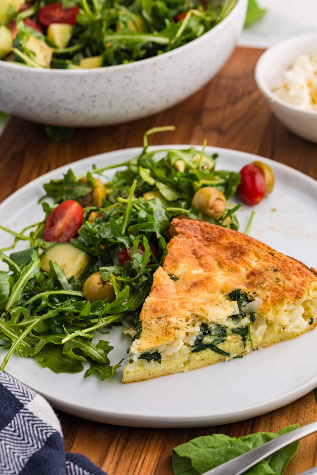 A slice of spinach and feta pie sits on a white plate beside an arugula salad with cherry tomatoes cucumber and green olives. A salad bowl and feta bowl in the background suggest an easy meal serving idea.