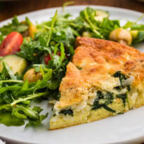 A slice of spinach and feta pie served on a white plate beside an arugula salad with cherry tomatoes cucumber and green olives. The browned top and creamy spinach filling make the dish look ready for lunch or dinner.
