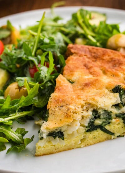 A slice of spinach and feta pie served on a white plate beside an arugula salad with cherry tomatoes cucumber and green olives. The browned top and creamy spinach filling make the dish look ready for lunch or dinner.