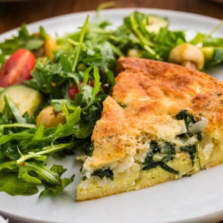 A slice of spinach and feta pie served on a white plate beside an arugula salad with cherry tomatoes cucumber and green olives. The browned top and creamy spinach filling make the dish look ready for lunch or dinner.
