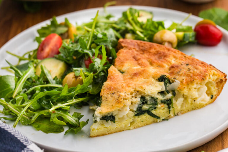 A slice of spinach and feta pie served on a white plate beside an arugula salad with cherry tomatoes cucumber and green olives. The browned top and creamy spinach filling make the dish look ready for lunch or dinner.