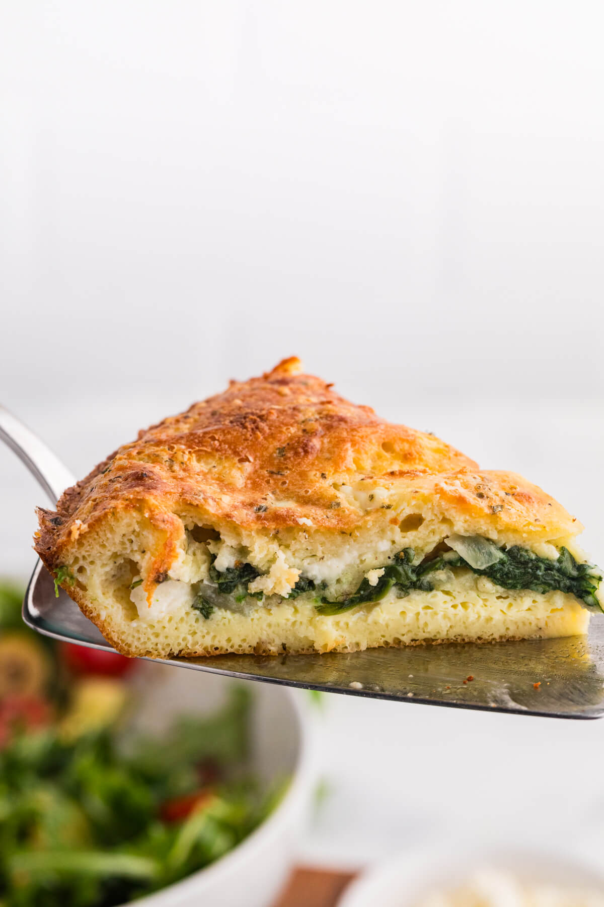 Close up slice of spinach and feta pie on a metal spatula with a golden browned top and a visible layer of spinach and cheese filling. A blurred green salad below suggests the slice is being served as part of a meal.