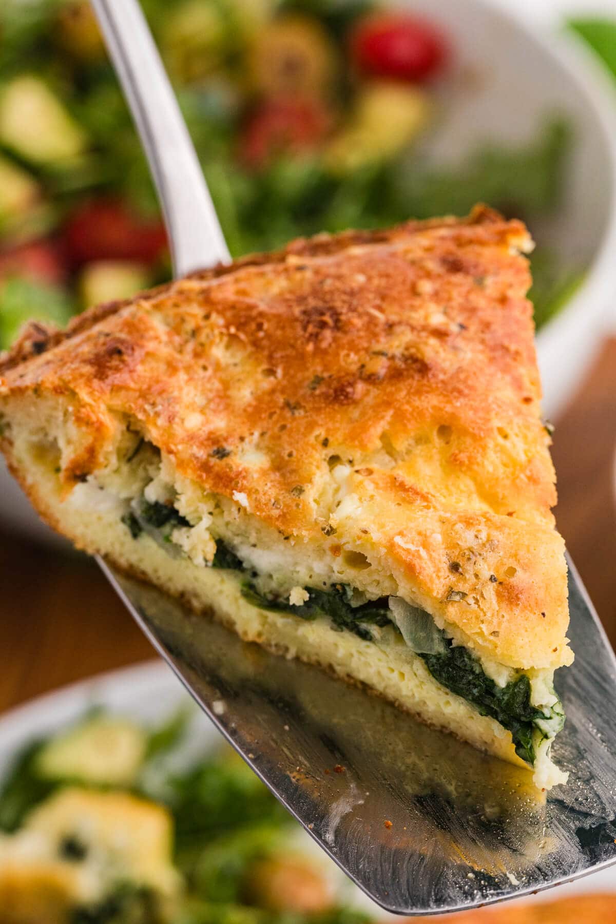 A slice of savory spinach and feta pie rests on a metal spatula with its golden browned top and green spinach filling visible. A bowl of salad is blurred in the background to show the pie ready to serve.