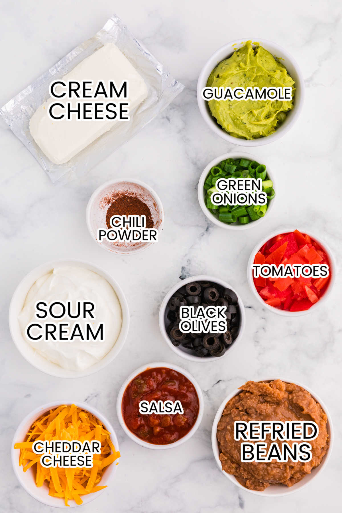 Ingredients for 7 Layer Dip, including refried beans, cream cheese, sour cream, guacamole, cheddar, salsa, tomatoes, black olives, green onions, and chili powder.