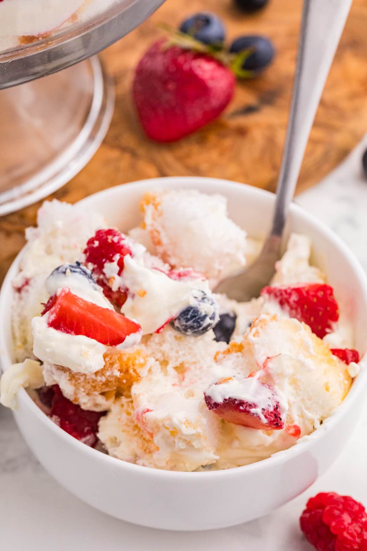 Serving of Angel Food Cake Trifle with fluffy cake, creamy vanilla filling, strawberries, blueberries, and raspberries in a white bowl.