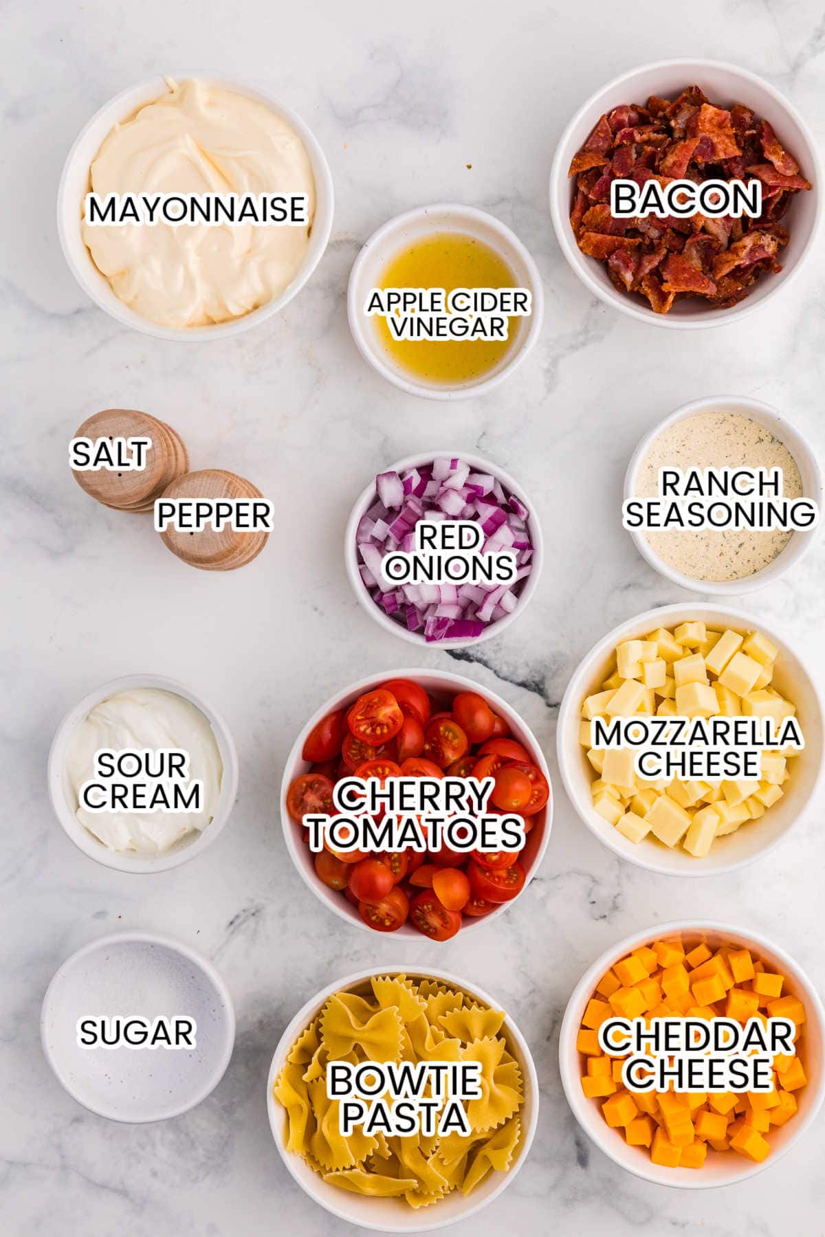 Ingredients for Million Dollar Pasta Salad arranged in bowls, including bowtie pasta, bacon, cheddar, mozzarella, cherry tomatoes, red onion, mayonnaise, sour cream, ranch seasoning, apple cider vinegar, sugar, salt, and pepper.