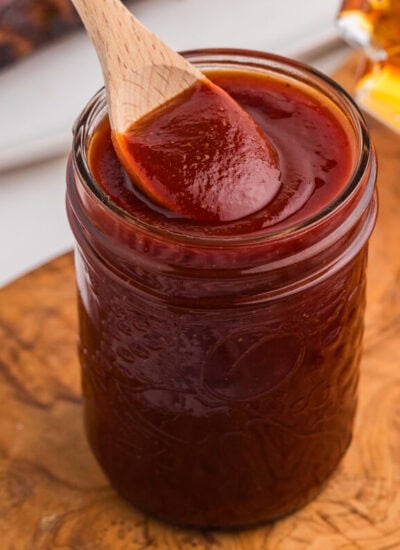 Homemade maple BBQ sauce in a mason jar with a wooden spoon, maple syrup, and glazed ribs nearby.