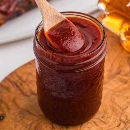 Homemade maple BBQ sauce in a mason jar with a wooden spoon, maple syrup, and glazed ribs nearby.