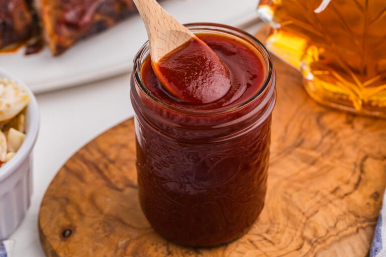 Homemade maple BBQ sauce in a mason jar with a wooden spoon, maple syrup, and glazed ribs nearby.