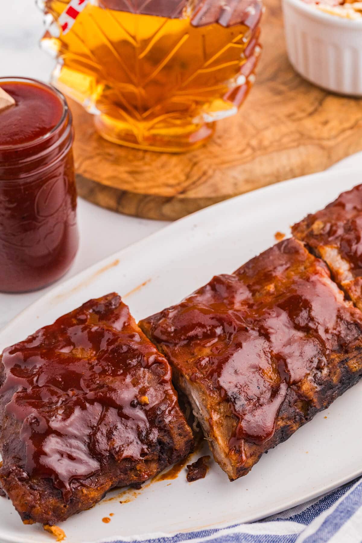 Maple BBQ sauce glazed ribs on a white platter with a jar of sauce and maple syrup nearby.
