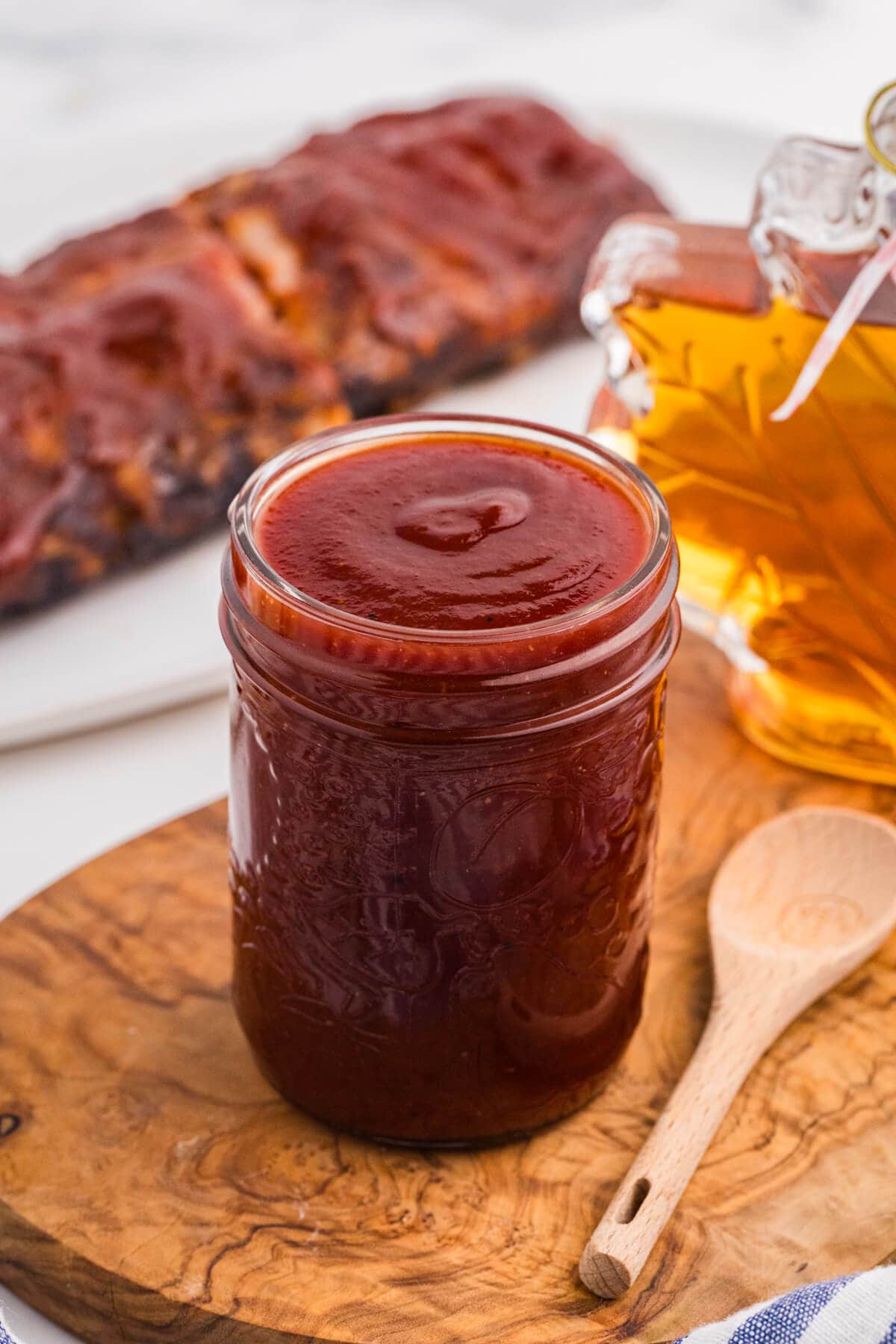 Homemade maple BBQ sauce in a mason jar beside a maple syrup bottle, with glazed ribs in the background.
