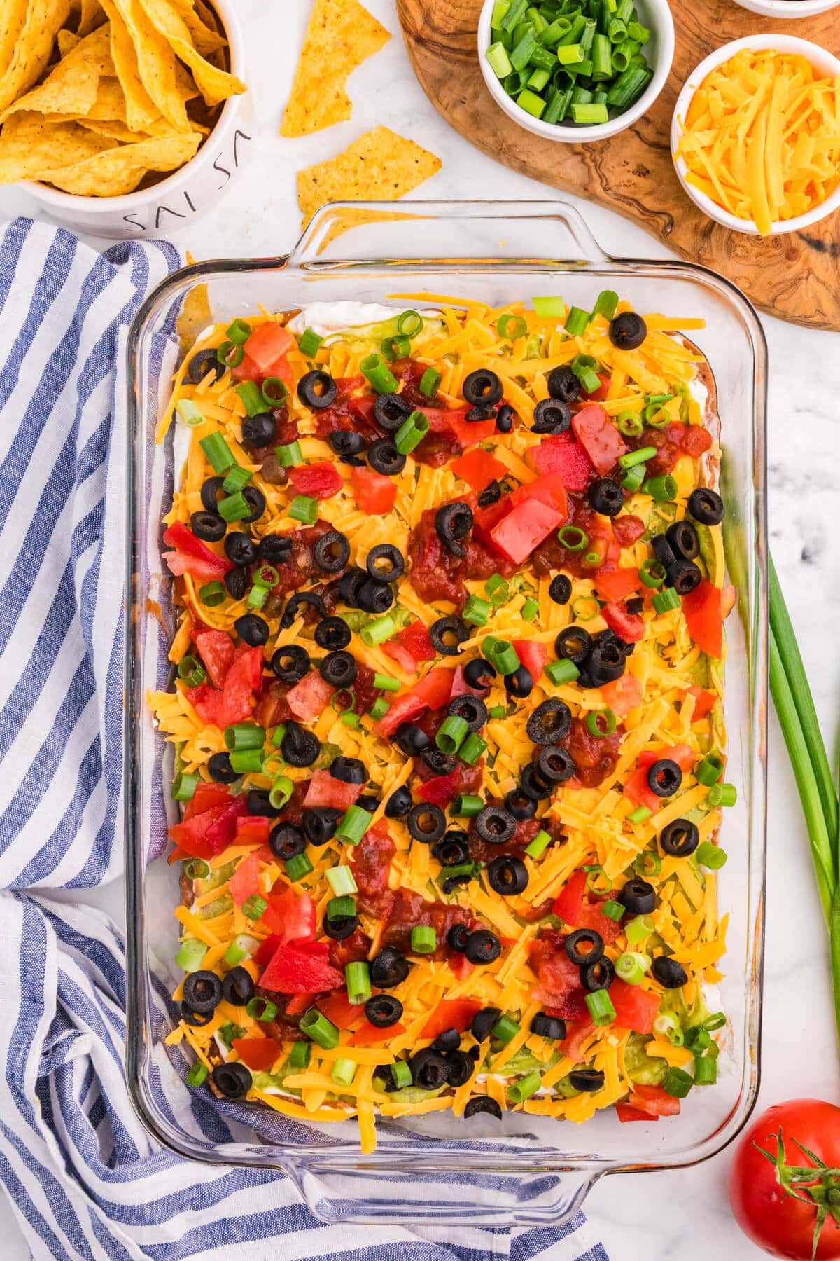 Overhead view of 7 Layer Dip topped with cheddar cheese, tomatoes, salsa, black olives, and green onions, served with tortilla chips.