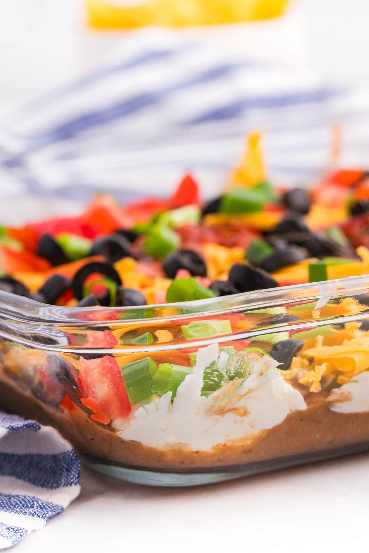 Side view of 7 Layer Dip in a glass baking dish, showing layers of refried beans, cream cheese mixture, guacamole, cheddar, tomatoes, black olives, and green onions.