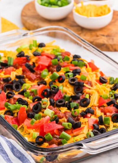 7 Layer Dip in a glass baking dish topped with cheddar cheese, tomatoes, salsa, black olives, and green onions, served with tortilla chips.