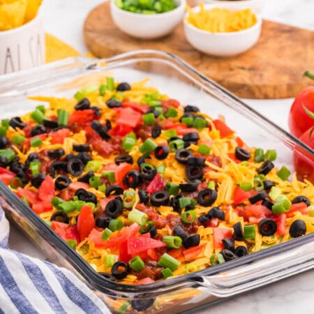 7 Layer Dip in a glass baking dish topped with cheddar cheese, tomatoes, salsa, black olives, and green onions, served with tortilla chips.
