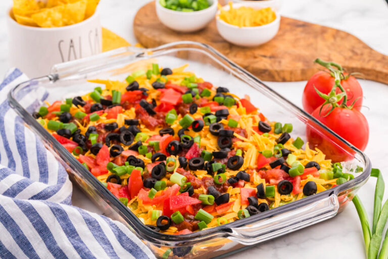 7 Layer Dip in a glass baking dish topped with cheddar cheese, tomatoes, salsa, black olives, and green onions, served with tortilla chips.