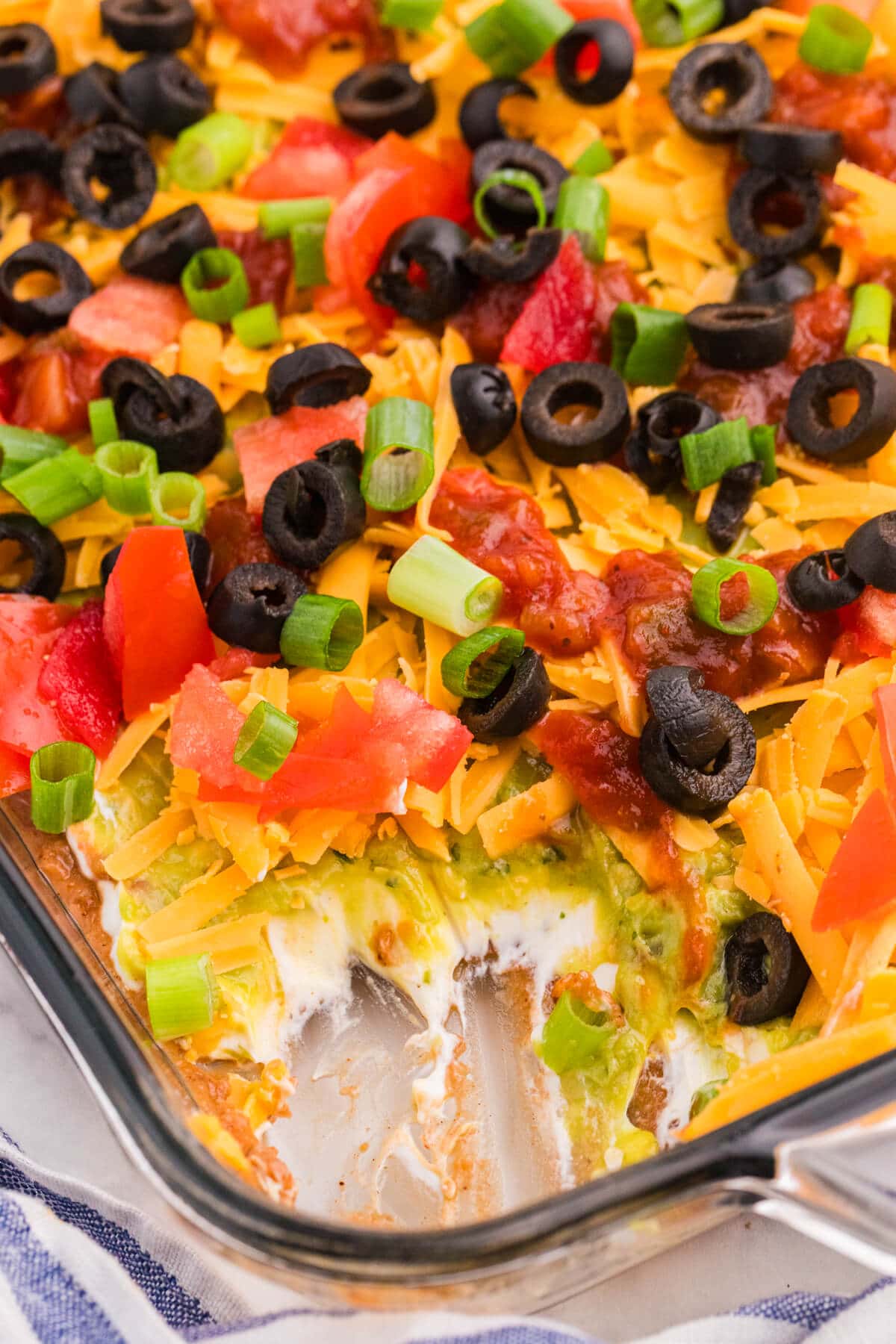 Close-up of 7 Layer Dip with a scoop removed, showing layers of refried beans, cream cheese, guacamole, cheddar, salsa, tomatoes, black olives, and green onions.